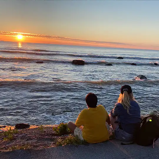 Två personer sitter vid vattnet med ryggen mot betraktaren och tittar på solnedgången.