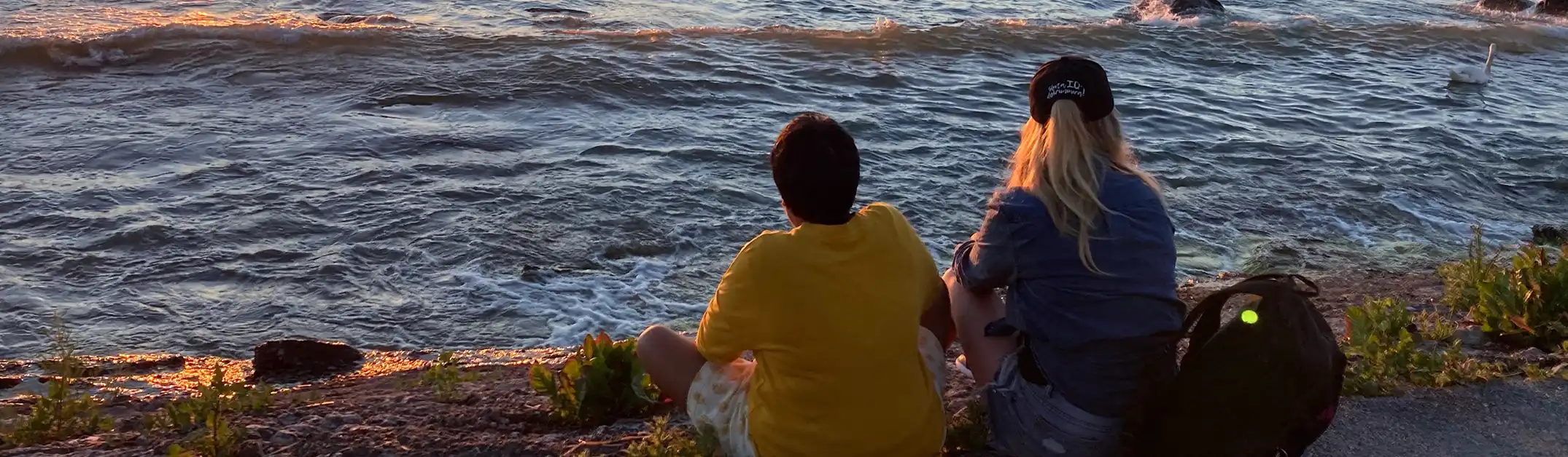 Två personer sitter vid vattnet med ryggen mot betraktaren och tittar på solnedgången.